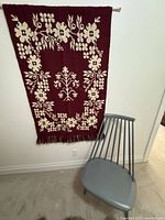 Front view of vintage Finnish Takana weaving wall hanging in dark maroon with cream floral patterns, showing full length and fringe bottom, hanging on wall next to a chair.