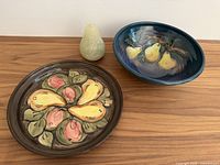 All three items together on wooden surface: a brown round floral platter, a blue fruit bowl with pears, and a pale green pear-shaped candle.