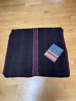 Top-down photo of folded dark woven wool manta with magenta and purple vertical embroidered strip along center and edges next to reference book on wooden floor.