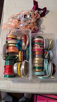 Two clear plastic bins filled with assorted ribbon spools of various colors, patterns, and textures, plus loose peach and pink sheer ribbons on top