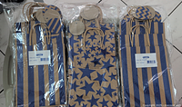 Three packages of kraft paper gift bags in two styles, blue star pattern and blue stripe pattern visible. Each bag has a blank kraft paper tag and handles, packaged and sealed in plastic.