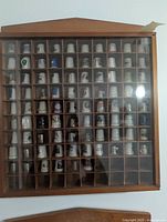 Full front view of the wooden display case mounted on the wall with 86 assorted thimbles in compartments behind plexiglass sliding cover.