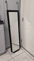 Full length black frame mirror standing against a door and cabinet, showing the overall condition and size relative to standard door.