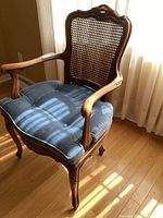 Front angle of the vintage Louis XV style wooden armchair with double cane backrest and blue tufted seat cushion, showing ornate carvings and armrest.