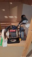 Ryobi BGH6110 bench grinder with buffing and grinding wheels, attached lamp, and power cord on wooden shelf.