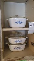 Three white CorningWare casserole dishes with Blue Cornflower pattern stacked inside a cupboard, each with a glass lid.