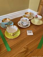Overall photo showing five vintage bone china cups and saucers of different colors and floral patterns placed on wooden floor.