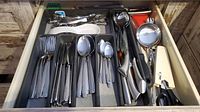 Drawer open showing organized stainless steel cutlery and kitchen utensils including forks, knives, spoons, ladle, tongs, and peeler.