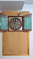 Open wooden cabinet with a mounted dart board inside, darts in a holder above, and chalk scoreboards on each door.