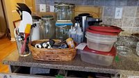 Wide view of kitchen countertop with jars, food storage containers, basket of kitchen tools, utensils in glass, and salad bowls