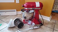 Red KitchenAid Artisan Plus stand mixer with stainless steel bowl and attachments laid out, including housing, drums, shield, spatula