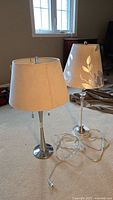Two table lamps standing on carpeted floor near window and chair, one with silver base and beige suede-like shade, other clear base with beige shade with leaf pattern cutouts