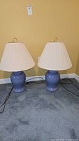 Two matching ceramic table lamps with blue bases and cream pleated fabric shades shown on carpeted floor against yellow wall.