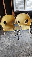 Two vintage yellow plastic wicker style patio chairs and one mosaic top round metal table shown from the front.