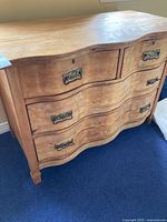 Front and right side view of the wooden dresser showing three drawers with ornate metal handles and wavy front paneling.