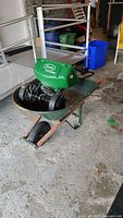 Wide angle photo showing the Scotts seed spreader placed inside the Garant wheelbarrow on a garage floor with various items in the background.