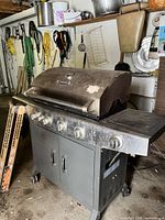 Four burner BBQ Tek gas grill in a garage setting with hanging tools and storage in background. Stainless steel lid and control panel with visible dirt and scuff marks.