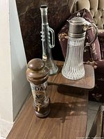 Three bar accessories on wooden surface: ribbed crystal pitcher with metal lid, narrow clear glass bottle with side handle, and wooden vodka dispenser with glass center section and brass spigot.