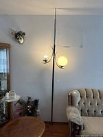Full view of black and chrome floor lamp positioned near wall and couch, showing two globe light holders with one bulb lit and one missing globe.