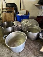 Overview photo showing large and medium aluminum pots, lids, and metal strainer arranged on garage floor.
