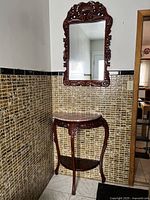 Front view of the carved cherry wood entrance table with marble top and lower shelf under an ornate wood framed mirror hung on tiled wall