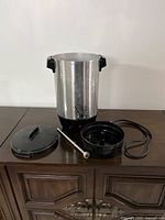30-cup coffee urn with all parts including lid, drip tray, rod, and sealing ring placed on a table, metal body with black handles.