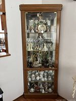 Front view of tall oak corner curio cabinet displaying porcelain figurines, glassware, and collectibles inside. Closed glass door with visible oak wood frame and brass hardware.