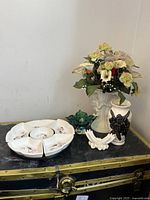 Cherub vase with artificial flowers, green and blue glass candy dish, ceramic plate with compartments, black and white ceramic vase and a small decorative ceramic wings holder placed on a black surface.