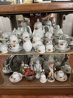 Display inside curio cabinet showing white porcelain tea set with floral designs and various pieces including teapot, coffee pot, sugar bowl, creamer, cups and saucers on glass shelf.