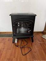 Front view of black Komodo electric fireplace standing on wooden floor with visible power cord.