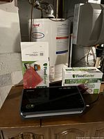 Foodsaver vacuum sealer machine on a wooden surface with unopened boxes of vacuum sealer bags behind it. Background shows part of basement setup.