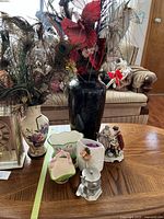 Photo showing tall black vase with artificial floral arrangements including peacock feathers, placed on wooden table near other ceramic and decorative items.