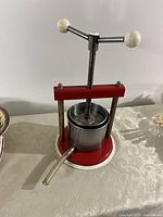 Front view of Italian Torchietto press showing red metal frame, screw press handle with white knobs, metal cylindrical basket, and juice outlet tube