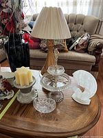 Photo showing wooden base lamp, candle holder with candles, 2-tier glass dessert dish, small candy dish, and white glass figurine arranged on wooden table in living room.