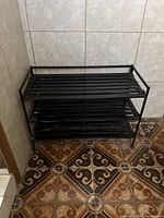 Front view of the black metal three-tier shoe rack placed against a tiled wall and floor.