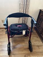 Front view of Nexus red collapsible walker with armrests, padded seat, and wheels, positioned indoors on wood floor.