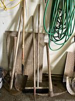 Photo showing various long-handled outdoor tools leaning against a wall including multiple shovels, a garden hoe, a push broom, and rakes, with a green garden hose coiled above them.