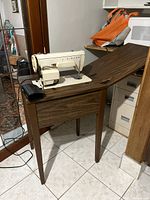 Singer sewing machine mounted on wooden cabinet with foot pedal and power cord visible.