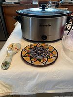 Bravetti slow cooker in stainless steel with glass lid, ceramic spoon holder and ceramic trivet with colorful design placed on a white tablecloth.