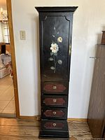 Front view of tall black wooden cabinet with floral hand-painted design, one door, and four drawers beneath it. Visible scratches and wear on surface.