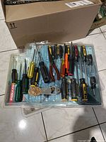 Top-down image of assorted screwdrivers, pliers, and metal accessories laid out in a plastic tray on floor. Tools mostly Fuller brand with yellow/black and red handles.