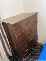 Front and side angled view of a medium brown wood veneer high boy dresser with four drawers. Shows scratches, scuffs, and wear marks with missing veneer edges on drawer fronts.