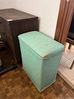 Side view of vintage mint green laundry basket placed beside a wooden chest of drawers, showing the texture and wear on the basket.