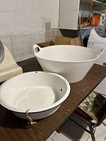 Two vintage metal wash basins on a table: one larger white basin with handles and black trim, showing wear and scratches; one smaller white basin with handle and rope tied to it.