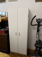 Closed white pantry cabinet standing upright next to exercise equipment and other furniture.