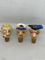 Three hand carved character bottle stoppers arranged side-by-side, showing detailed painting and carving.