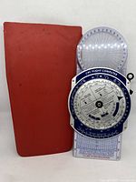 Front view of the Cessna CPC Flight Computer next to red protective case showing the mechanical dials and geometric scales.