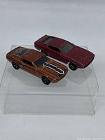 Photo showing both 2010 Hot Wheels '67 Shelby GT500 and 1970 Ford Mustang Mach 1 models positioned on a clear display stand, angled to show top and side details, highlighting color and condition.