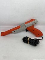 Front and side view of Nintendo NES Zapper light gun, showing orange and gray plastic body, trigger, and bundled cable.