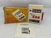 Photo showing the handheld slot machine, original yellow box, and instruction manual laid out on a surface.